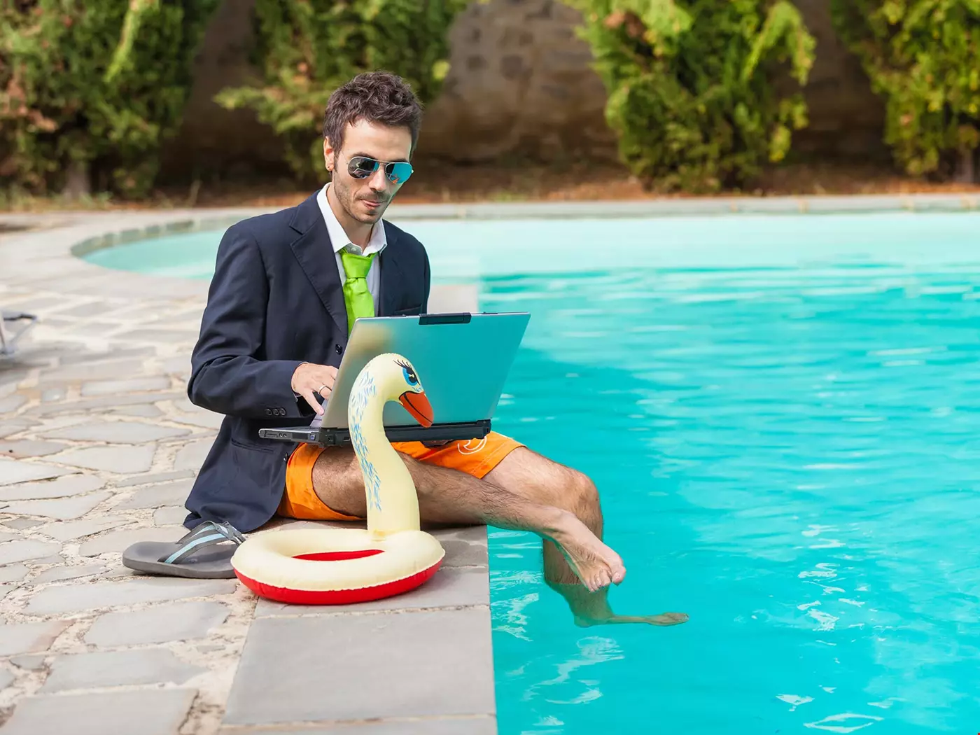 En man i kostym sitter med dator i knäet vid en pool, med fötterna ner i vattnet. Ser ut som ledighet, men jobbar