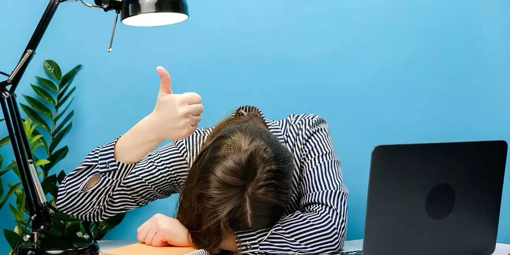 En ledsen medarbetare vid sitt skrivbord gömmer huvudet i händerna men håller samtidigt upp en tumme i luften som ett godkännande Akavia Aspekt
