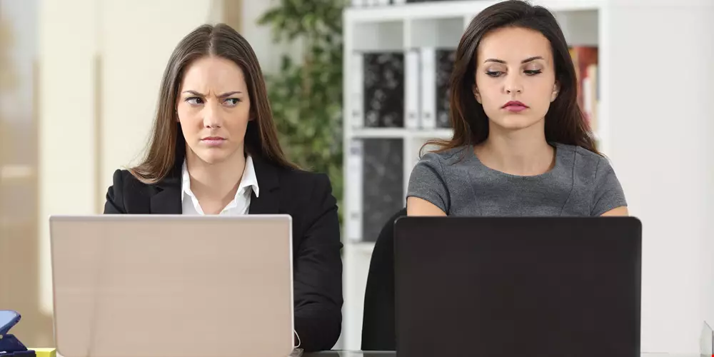 Två kvinnliga kollegor bredvid varandra på kontoret, ser mycket missnöjda ut med varandra, är troligen avundsjuka Akavia Aspekt