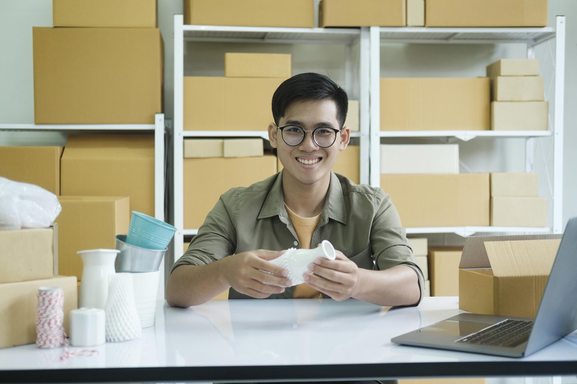 Ung man med glasögon sitter i lagermiljö och packar in beställningar i lådor har dator bredvid sig Akavia Aspekt
