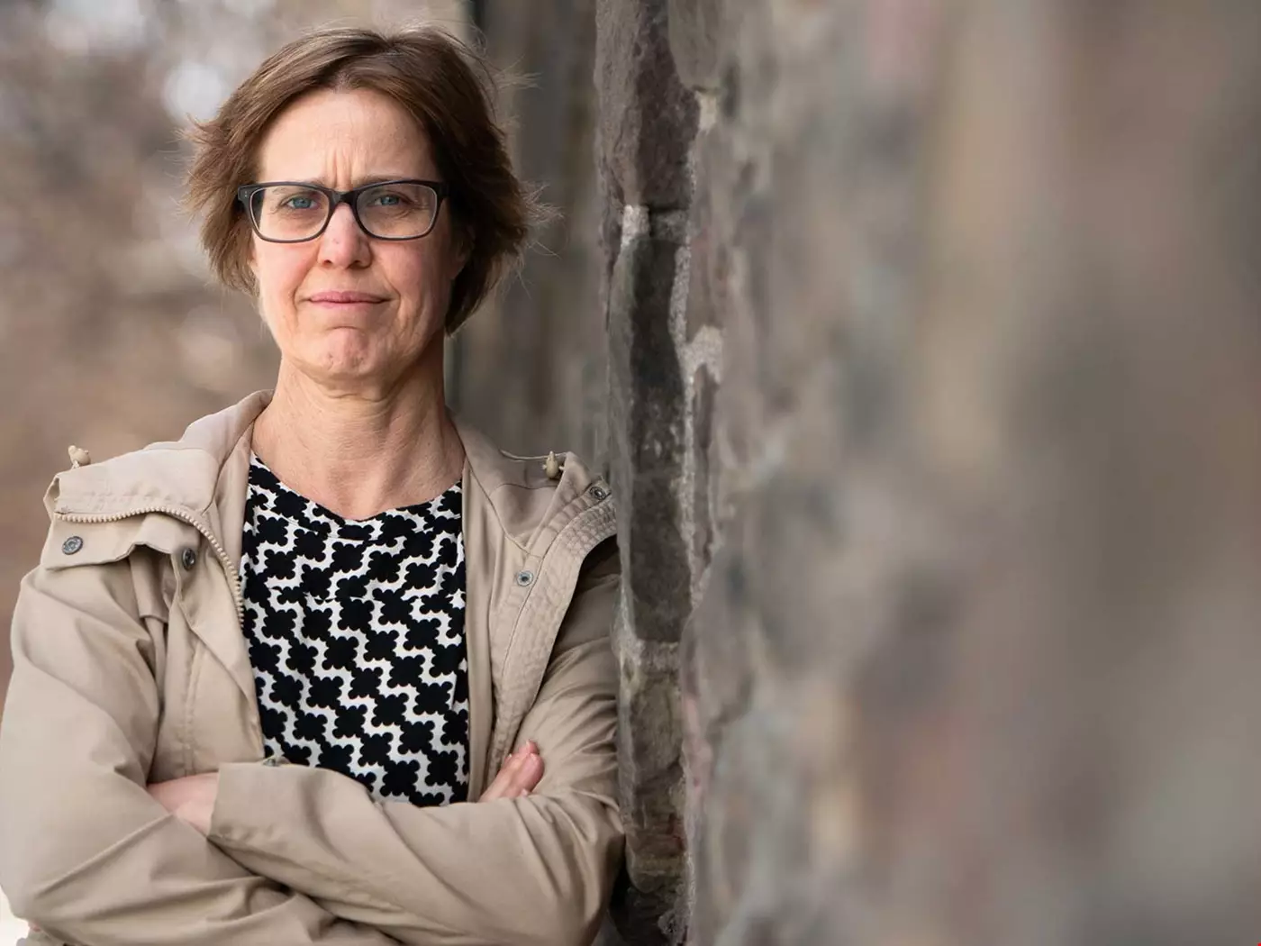 Kvinna, kort hår, glasögon, lutar sig mot en vägg, ljusa kläder