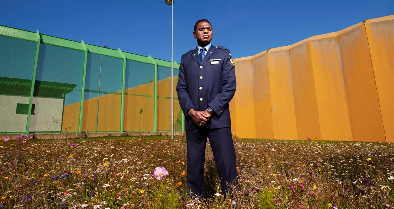 Medelålders man med uniform, står på en blommande sommaräng framför en flera meter hög gul cementmur samt en gallermur som omger ett fängelse