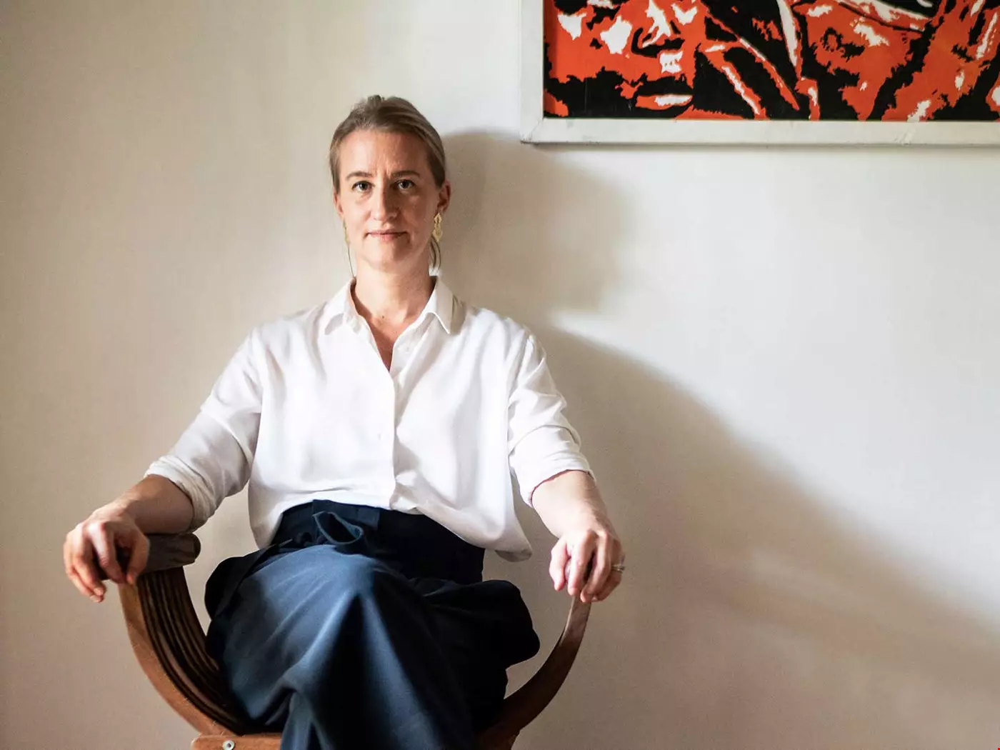 Kvinna med håret i hästsvans, ljus blus och mörka byxor, sitter i fåtölj mot ljus vägg