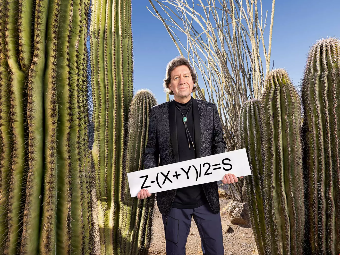 Porträtt Martin Dufwenberg i kavaj bland kaktusar i Arizona-öknen håller i skylt som säger Z-(X+Y)/2=S